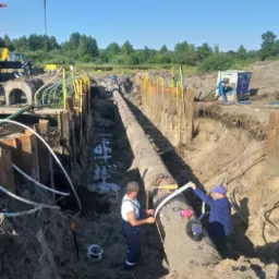 Dwaj pracownicy mierzą rurę ułożoną w głębokim wykopie zabezpieczonym deskami, w tle widoczny dźwig i maszyny budowlane.