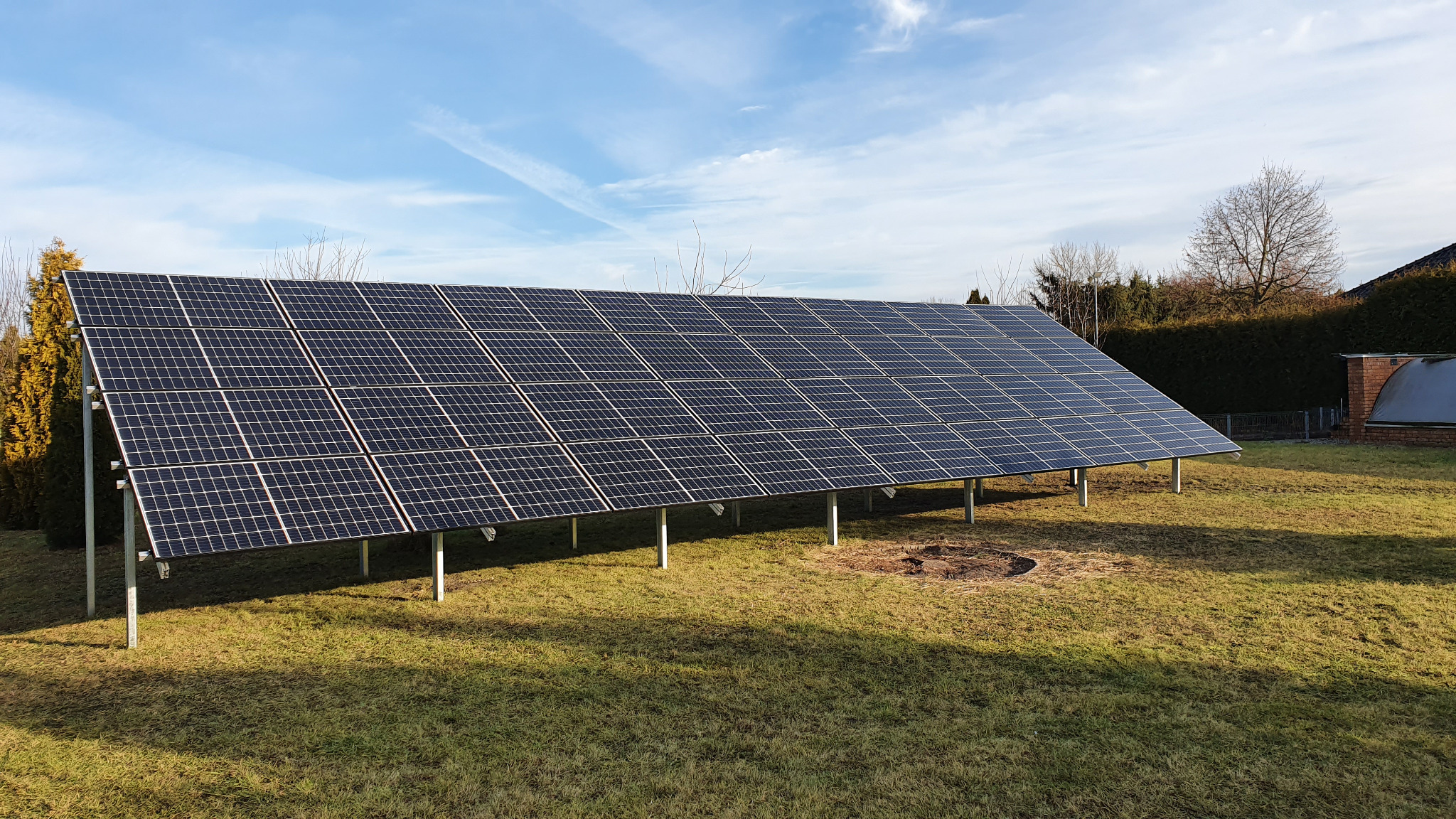 Instalacja fotowoltaiczna na metalowych podporach, umieszczona na trawniku w ogrodzie z widocznymi drzewami i niebieskim niebem z delikatnymi smugami chmur w tle.