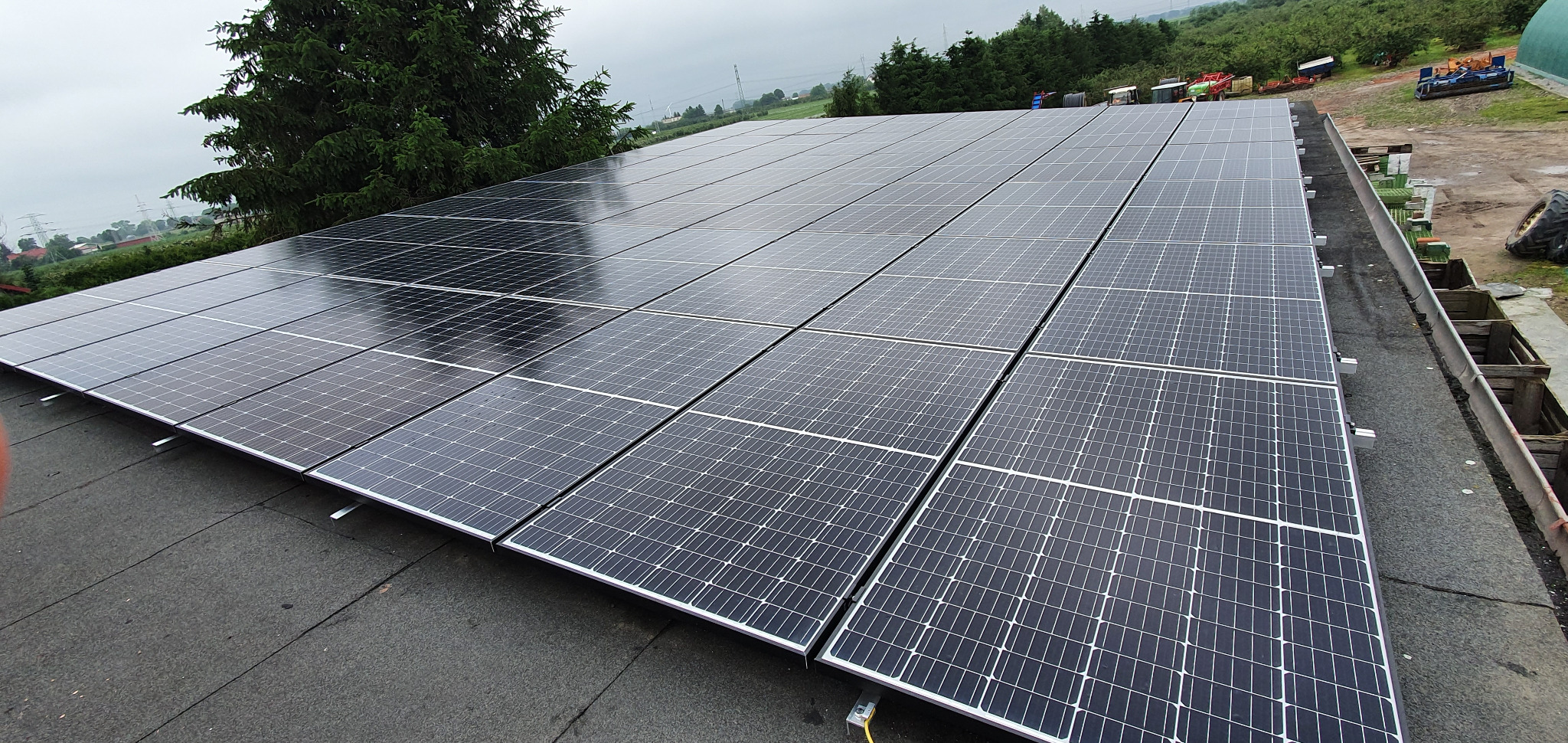 Rozległa instalacja fotowoltaiczna na dachu budynku gospodarczego, widok z góry, w tle zieleń i pochmurne niebo.