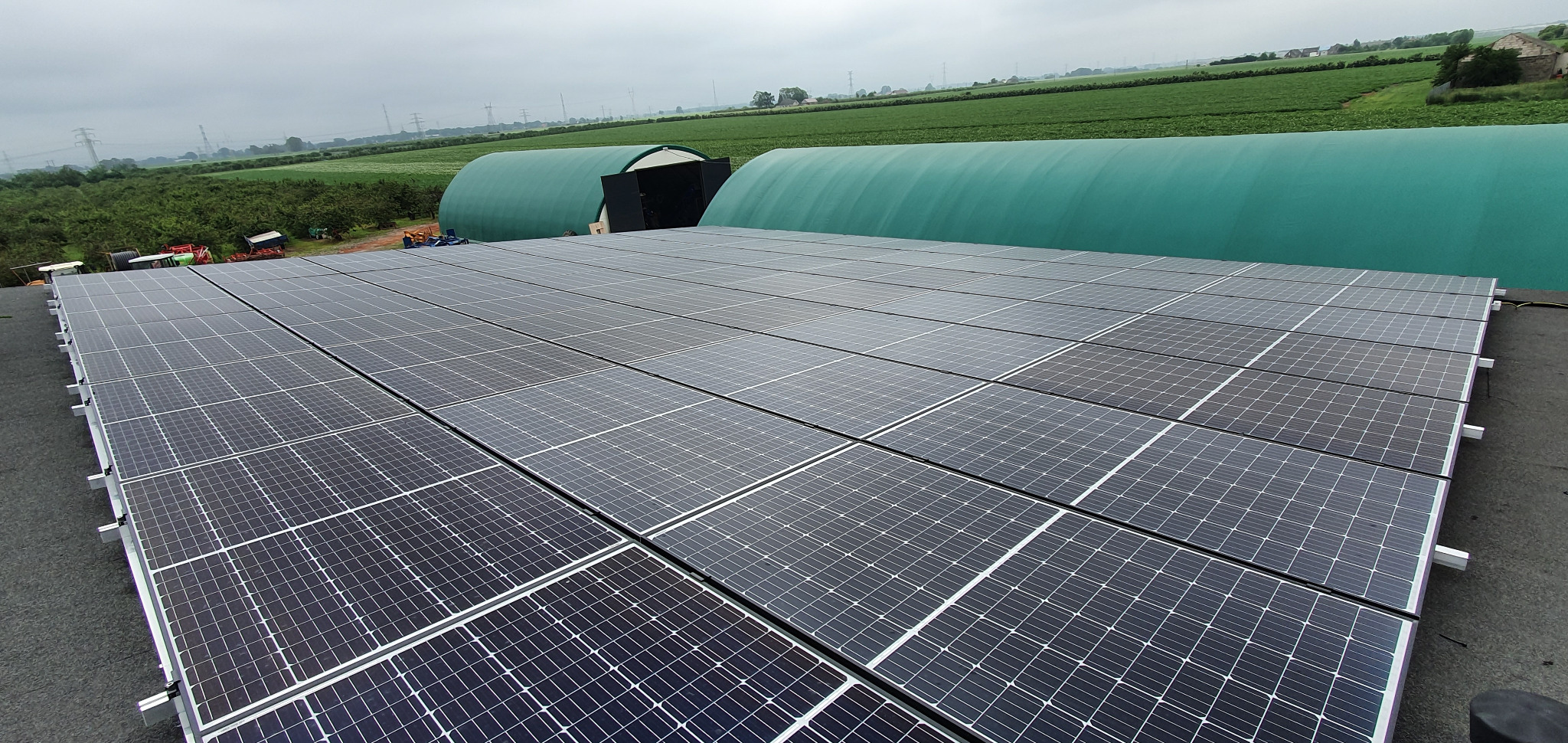 Rozległa instalacja fotowoltaiczna na dachu z widokiem na zielone tunele foliowe i odległe pola uprawne pod pochmurnym niebem.