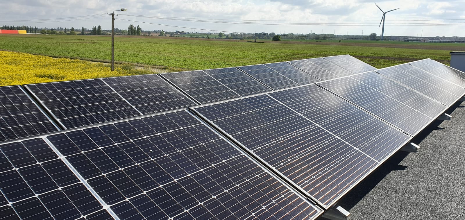 Rząd paneli słonecznych na dachu z widokiem na pola uprawne i turbinę wiatrową w oddali.