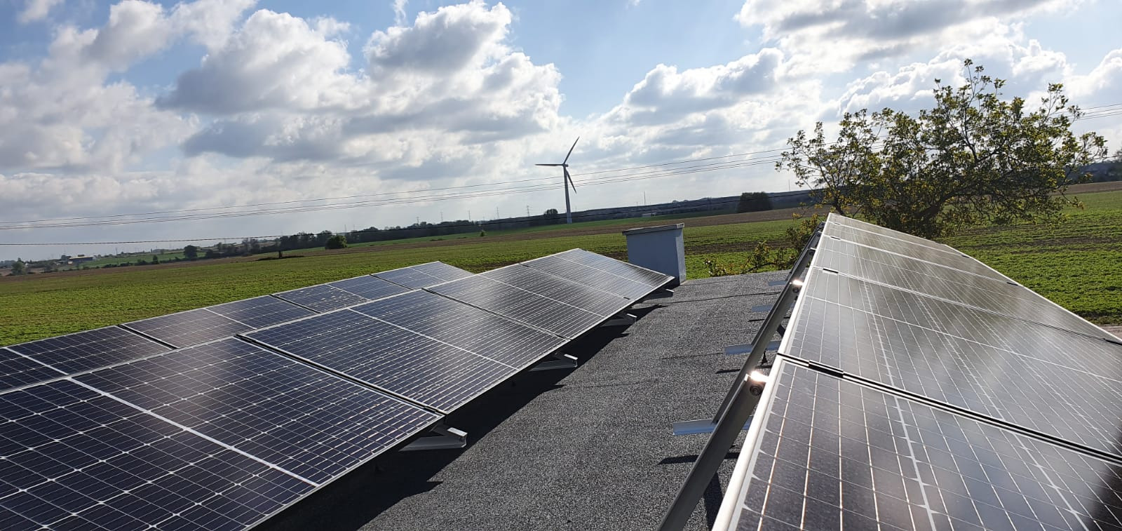 Instalacja paneli słonecznych na dachu z widokiem na pole uprawne i turbinę wiatrową w oddali, przy słonecznej pogodzie z chmurami.