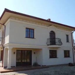 Dwukondygnacyjny dom murowany z beżową elewacją, ciemnobrązową stolarką okienną i balkonami z ozdobnymi, kutymi balustradami, widziany z perspektywy lekko z dołu.