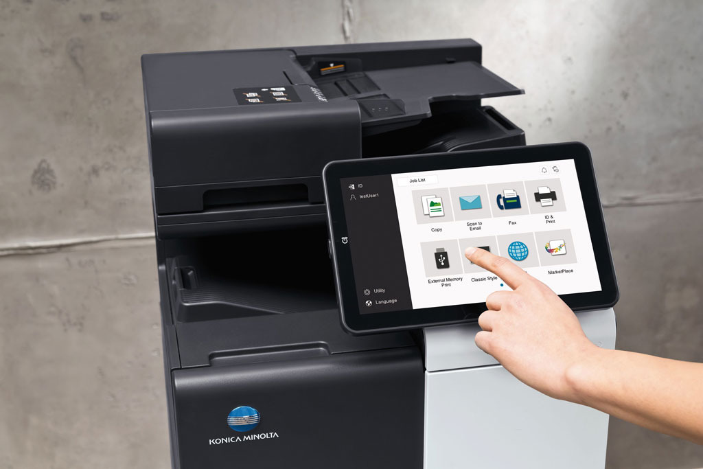 Czarno-biała kserokopiarka Konica Minolta z dotykowym panelem sterowania, na którym palec wskazuje ikonę 'Classic Style'.