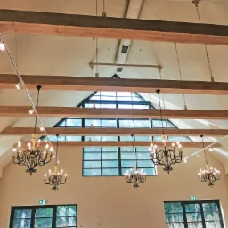 Wysokie wnętrze hali z drewnianymi belkami stropowymi, oświetleniem szynowym i wiszącymi żyrandolami, widoczne okna i drzwi z czarnymi ramami.