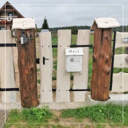 Drewniana furtka z jasnych desek i ciemnych bali, z metalową skrzynką na listy, na tle domu z czerwonymi kwiatami w oknach.