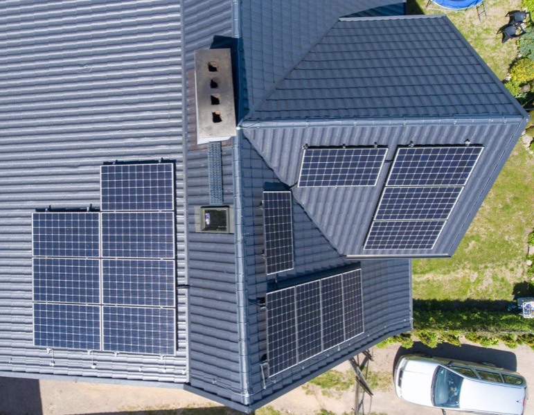 Widok z góry na dach domu z zainstalowanymi panelami fotowoltaicznymi w różnych konfiguracjach, obok komina i wentylacji, z samochodem zaparkowanym w pobliżu.