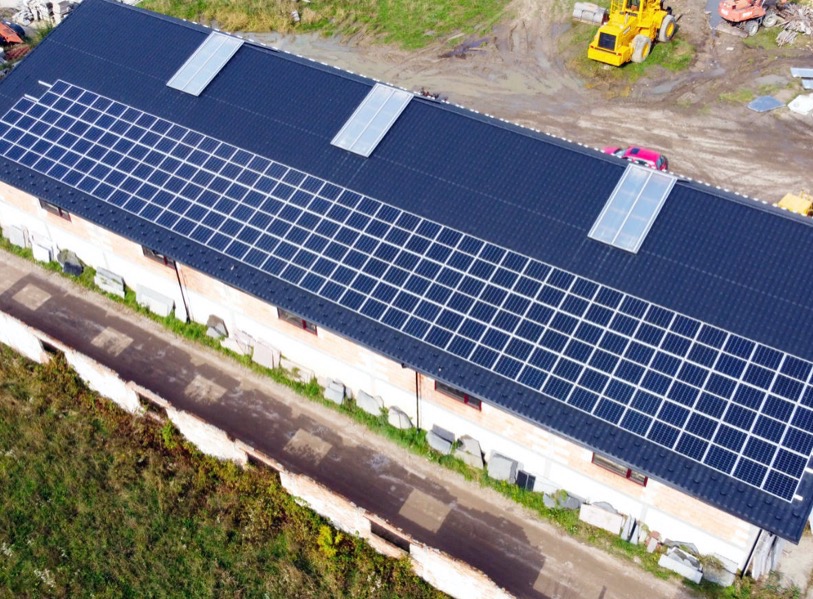 Widok z góry na budynek z cegły z zainstalowanymi panelami słonecznymi na dachu; na dachu widoczne również świetliki; w tle żółta koparka.