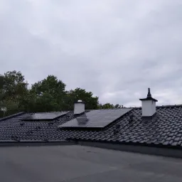 Czarny dach z dachówki ceramicznej z zamontowanymi panelami fotowoltaicznymi i kominami, widok z dołu na tle pochmurnego nieba i drzew.