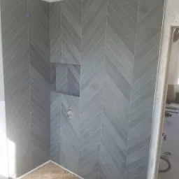 Szare płytki ułożone w jodełkę na ścianach kabiny prysznicowej z wnęką, widoczna instalacja wodna i zabezpieczone podłoże.