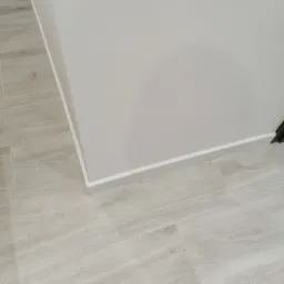 Ułożone jasne panele podłogowe imitujące drewno, przy białej ścianie z listwą przypodłogową.