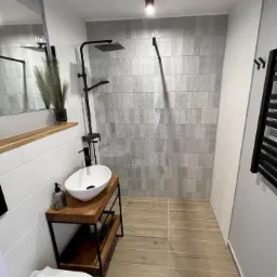 Nowoczesna łazienka z kabiną prysznicową typu walk-in, umywalką nablatową na drewnianym regale i grzejnikiem drabinkowym w kolorze czarnym.