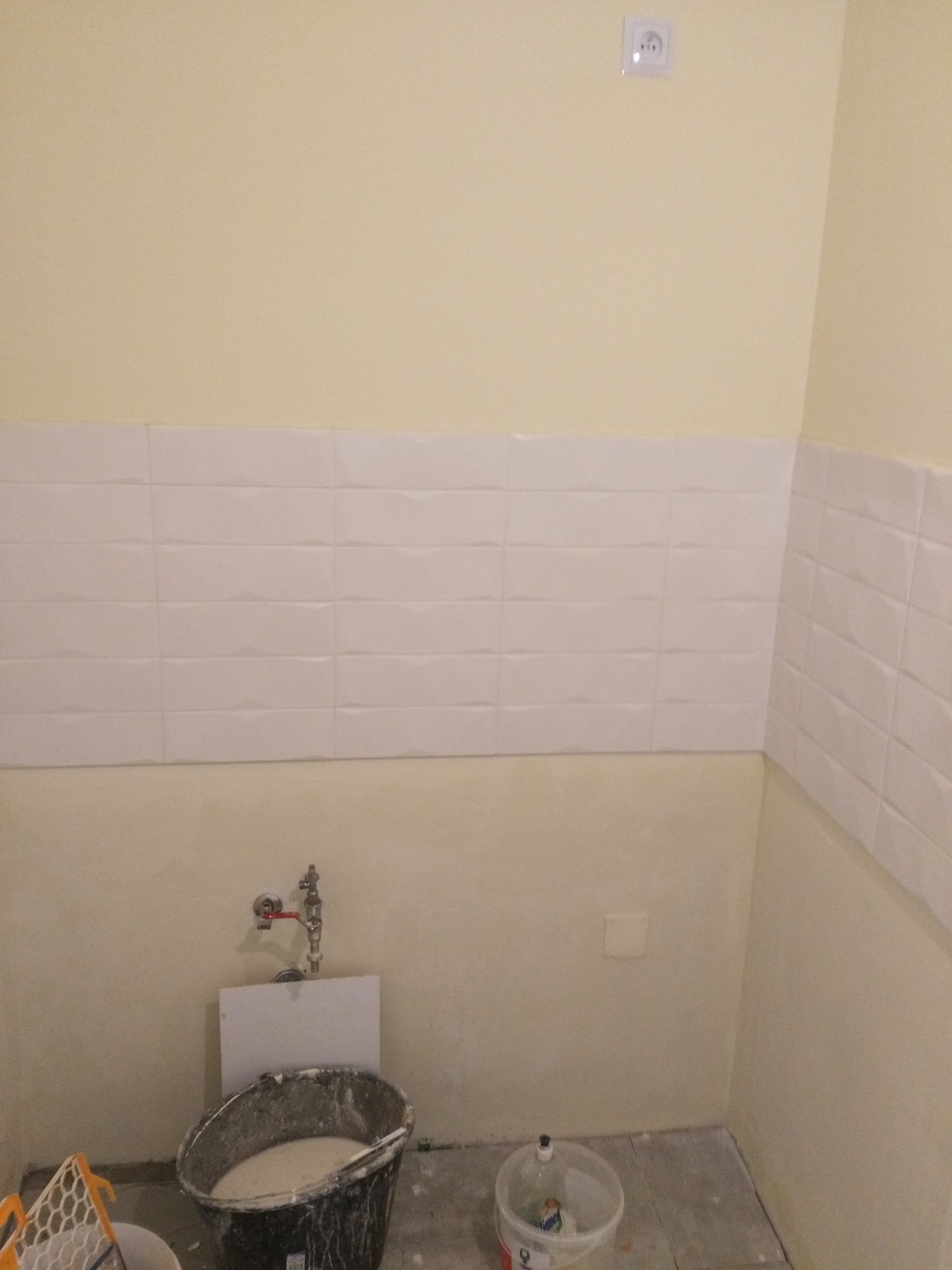 Ściana łazienki z ułożonymi białymi płytkami o trójwymiarowej fakturze, widoczne przygotowania do dalszych prac wykończeniowych: wiadro z zaprawą, pędzle, oraz odsłonięta instalacja wodna.