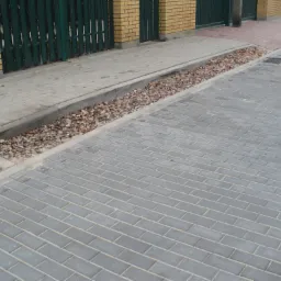 Szara kostka brukowa ułożona wzdłuż ulicy, z widocznym korytkiem wypełnionym kamieniami i liśćmi, oddzielającym chodnik od jezdni.