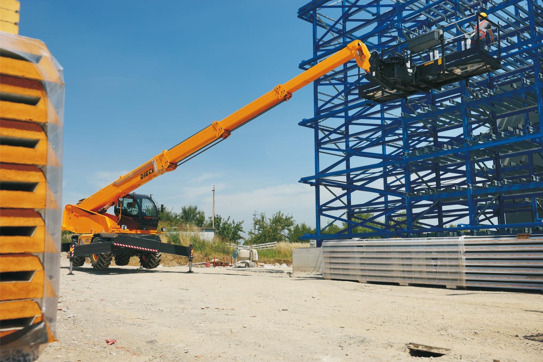 Żółta ładowarka teleskopowa Dieci Pegasus 40.16 z operatorem w koszu, na tle niebieskiej konstrukcji stalowej budynku w trakcie montażu, obok paleta z panelami warstwowymi.