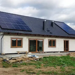 Niedokończony dom jednorodzinny z szarego bloczka z zamontowanymi panelami słonecznymi na dachu pokrytym ciemną dachówką, widoczne drewniane ramy okienne i drzwiowe, oraz stos desek na ziemi.