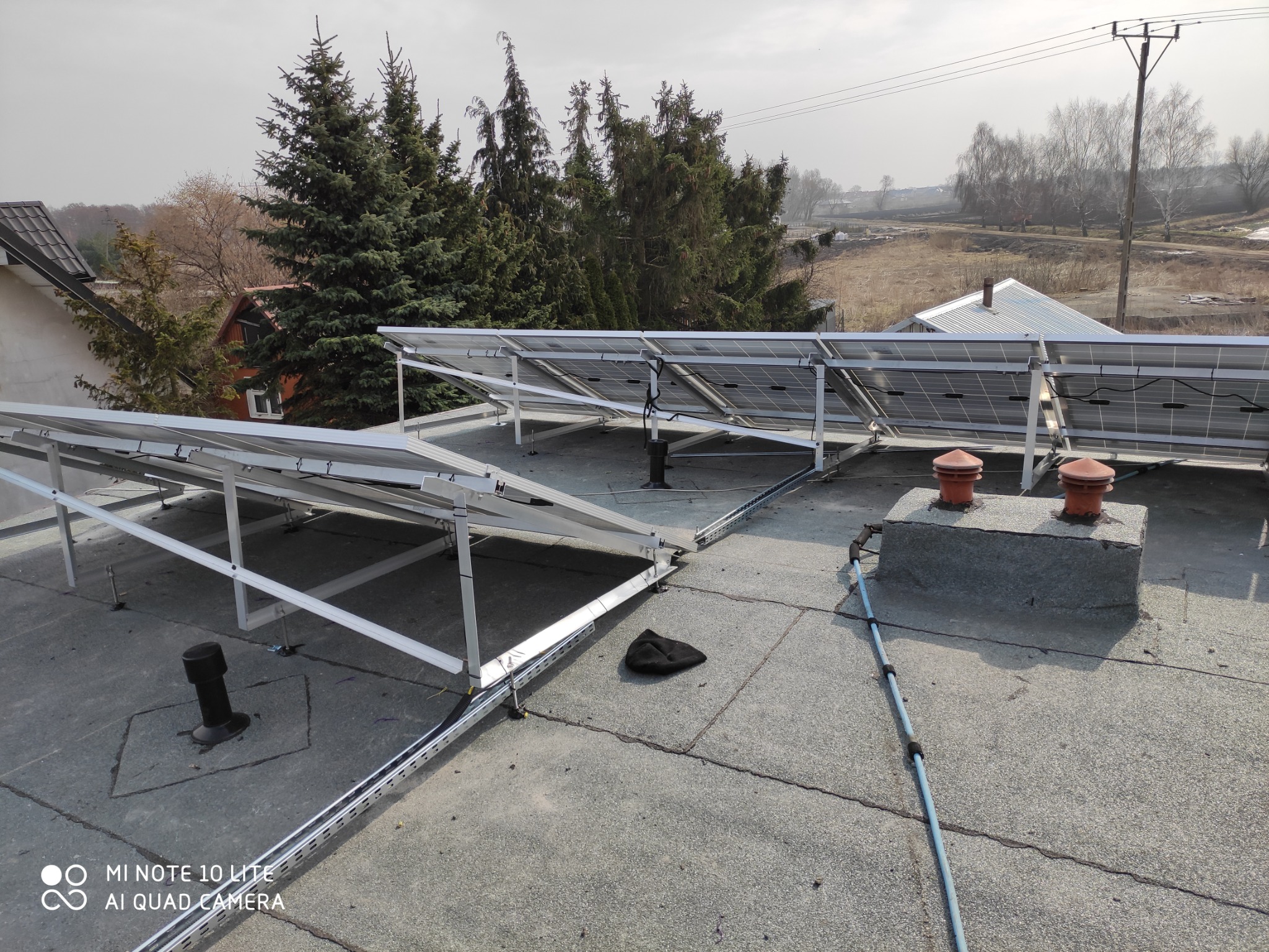 Panele słoneczne zamontowane na płaskim dachu z widocznymi elementami mocującymi, kominem wentylacyjnym i elementami instalacji elektrycznej w tle z drzewami i zabudowaniami.