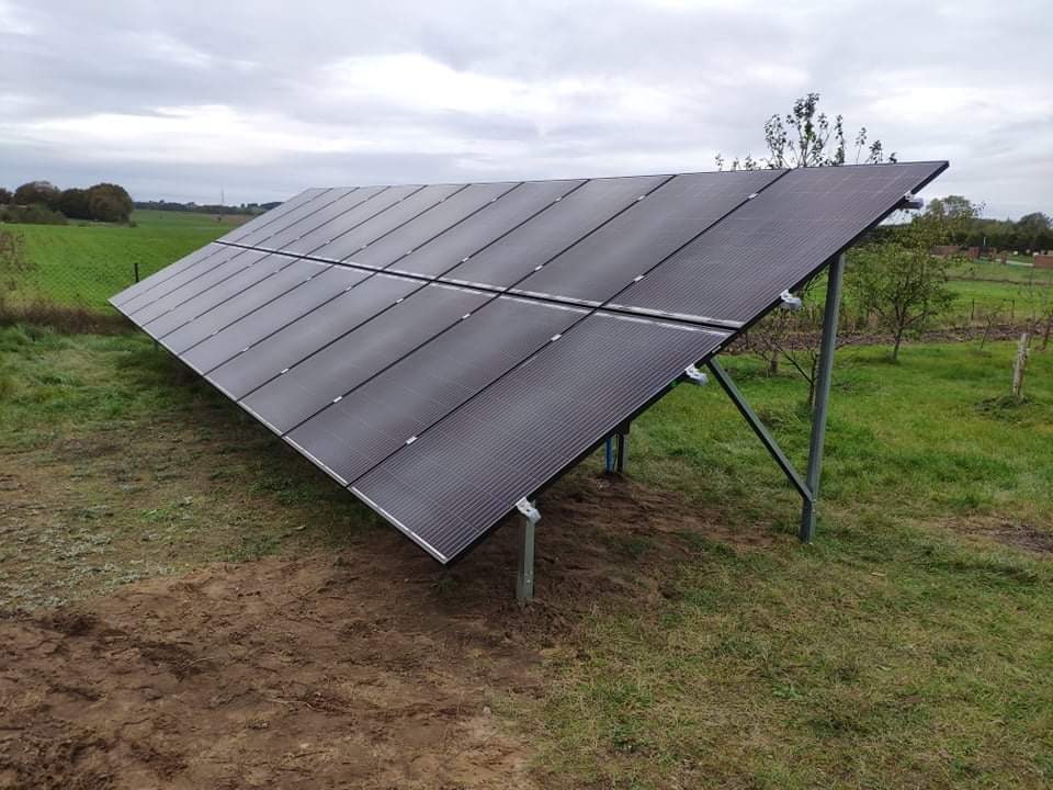 Rząd paneli słonecznych zamontowanych na metalowej konstrukcji na polu, widoczne łączenia między panelami, w tle zielone pola i pochmurne niebo.