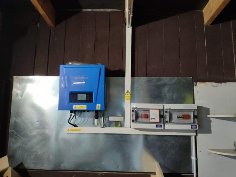 Niebieski falownik zamontowany na metalowej płycie wewnątrz budynku, obok skrzynki z zabezpieczeniami fotowoltaicznymi, na tle brązowej boazerii.