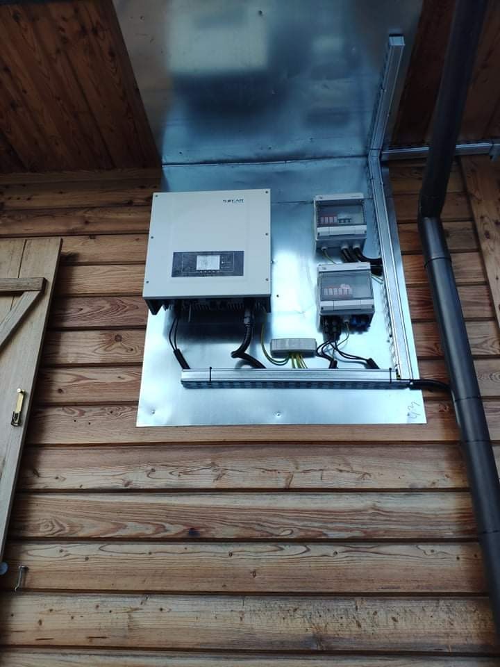 Zamontowany inwerter fotowoltaiczny z rozdzielnicą, na metalowej płycie przymocowanej do drewnianej ściany budynku gospodarczego, widoczne kable i rury instalacyjne.