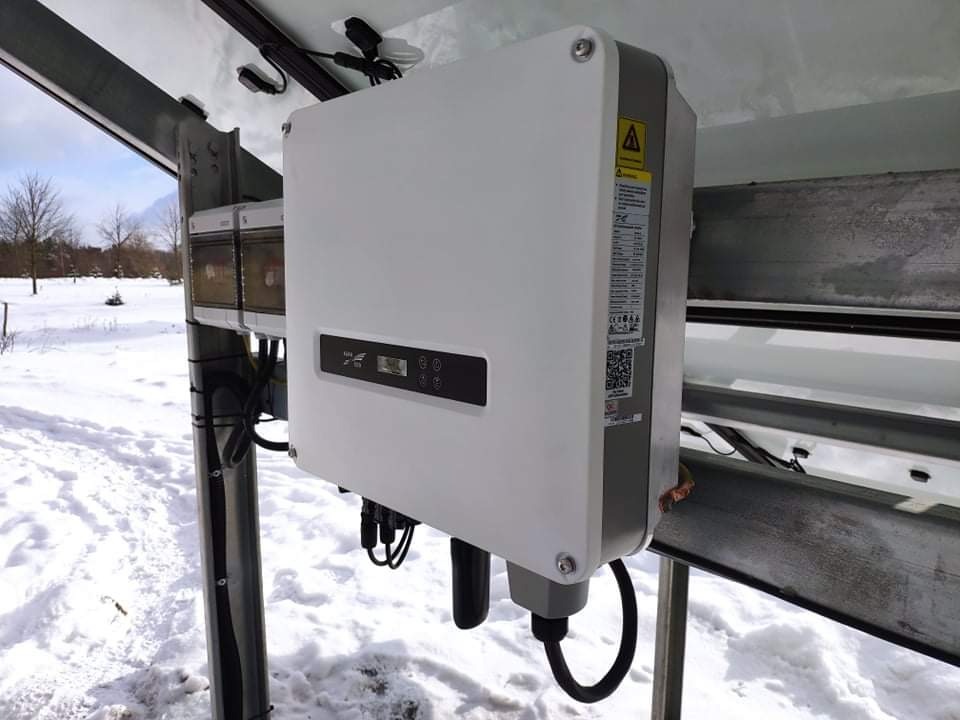 Biały inwerter zamontowany na metalowej konstrukcji w zasypanym śniegiem otoczeniu, widoczne kable i etykiety informacyjne.