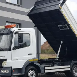 Biała wywrotka Mercedes Atego 1218 z podniesioną czarną skrzynią ładunkową, widok z boku na tle szarego budynku z poziomymi pasami.