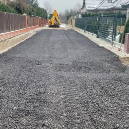 Świeżo ułożona, czarna nawierzchnia asfaltowa na drodze osiedlowej, z żółtą koparką JCB w tle i ogrodzeniami posesji po obu stronach.