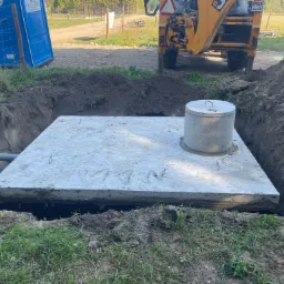 Betonowa płyta z włazem umieszczona w wykopie, widoczny fragment koparki i toaleta przenośna w tle.