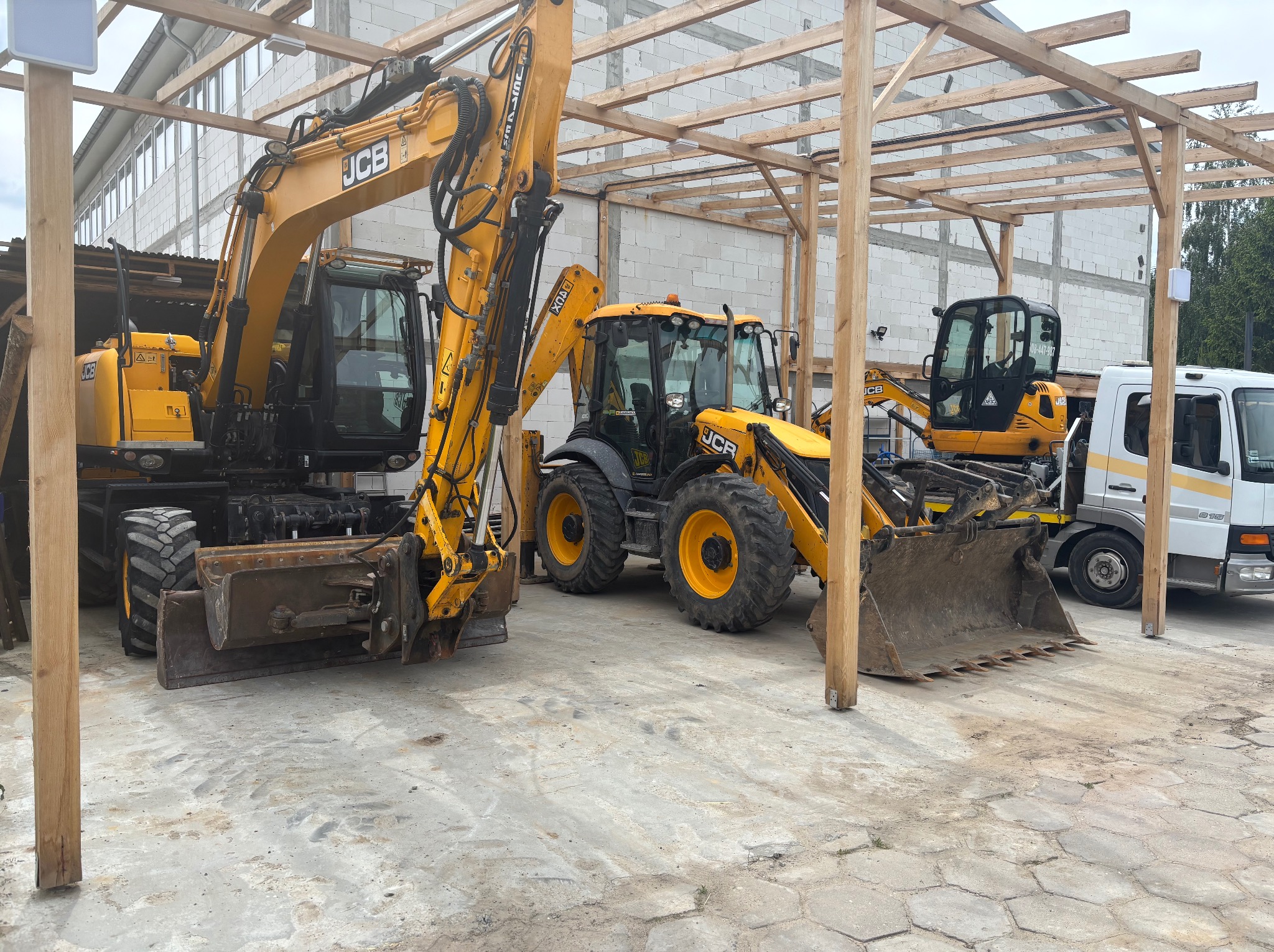 Żółte koparki JCB, w tym kołowa i koparko-ładowarka, stoją na placu budowy pod drewnianą konstrukcją. W tle widoczny budynek z białego bloczka i częściowo biała ciężarówka.