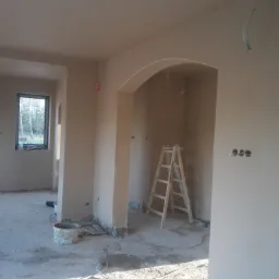 Wnętrze domu w Przemyślu po nałożeniu tynków, widoczny fragment ściany z wykończonym łukiem, drabina oparta o ścianę, oraz wiadro i narzędzia na podłodze.