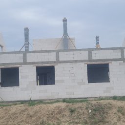 BUILDING PL - Wznoszony dom z białych bloczków, widoczne otwory okienne i kominy, pracownik wyrównuje teren przed budynkiem.