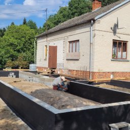 BUILDING PL - Pracownik zabezpiecza czarną izolacją przeciwwilgociową betonowe ściany fundamentów domu jednorodzinnego, widoczny fragment elewacji budynku z cegły.
