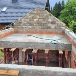 Budowa domu w trakcie realizacji: wylany betonowy strop, ściany z czerwonej cegły, widoczne podpory i szalunki, fragment murowanej ściany szczytowej z bloczków betonowych, zielony wąż na stropie.