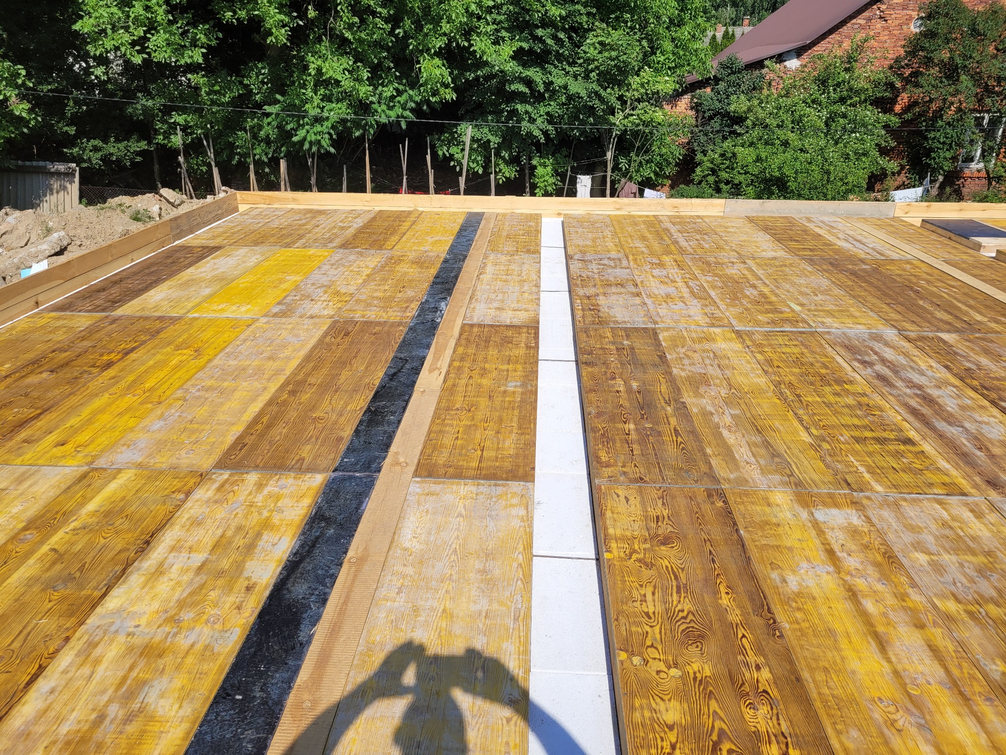 Przygotowany szalunek fundamentów z deskami w odcieniach brązu i żółci, z widocznym zbrojeniem i izolacją pionową, w tle zieleń drzew i fragment budynku.