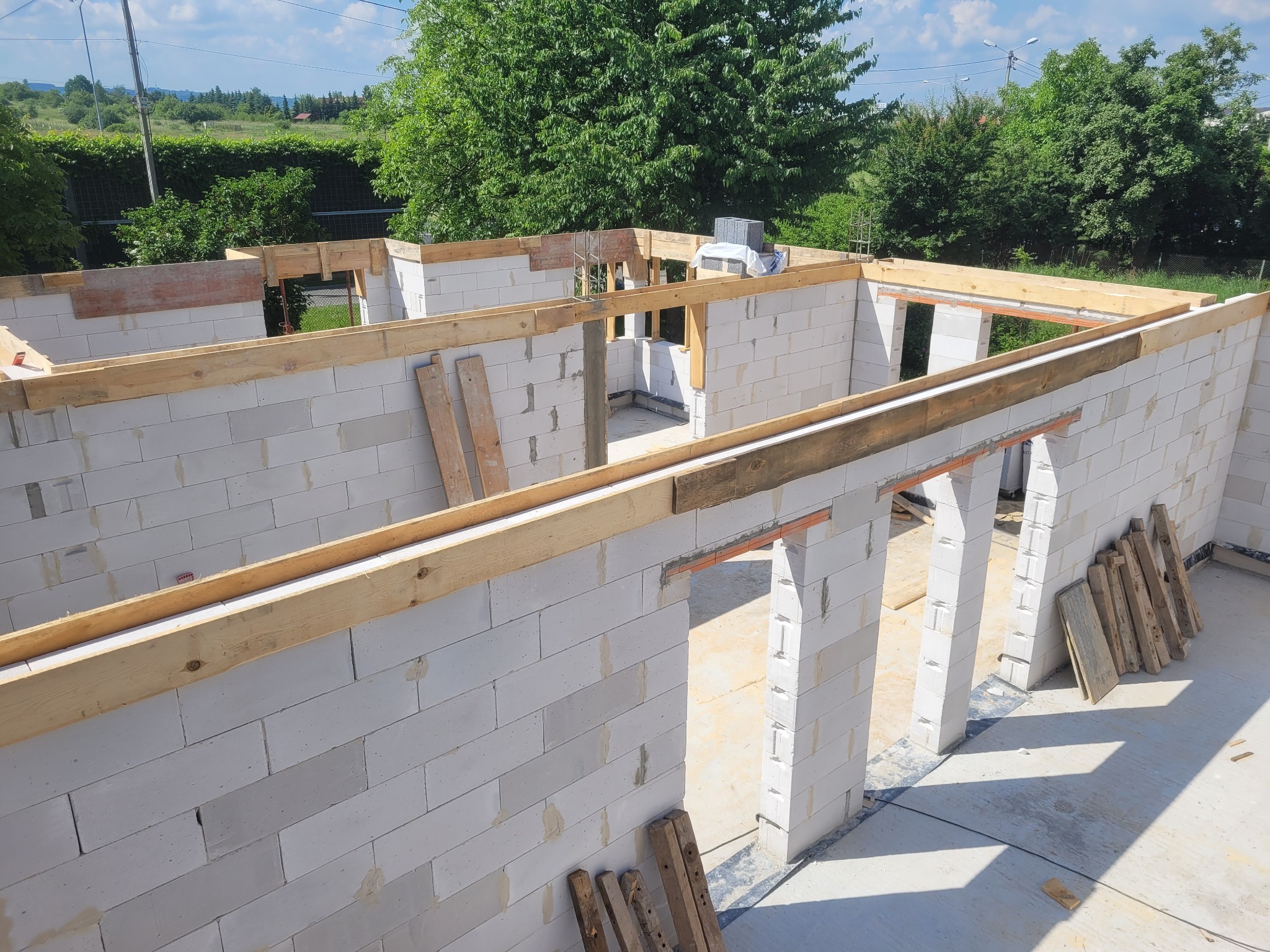 Wznoszenie ścian budynku z bloczków betonu komórkowego, widoczne drewniane belki stropowe i otwory okienne, w tle zieleń drzew i błękitne niebo.