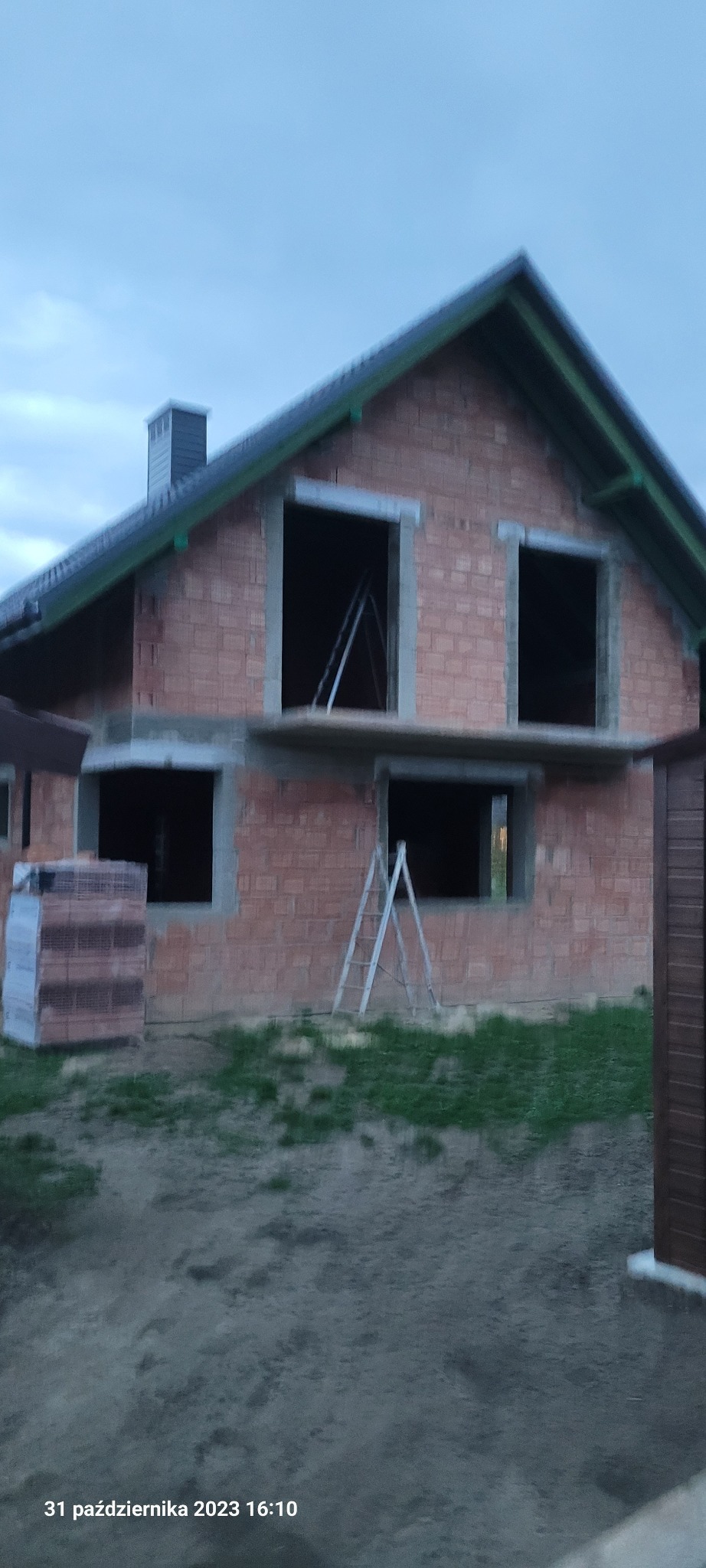 Surowy stan domu murowanego z czerwonej cegły z widocznymi otworami okiennymi, schodami i paletą cegieł na zewnątrz, w pochmurny dzień.