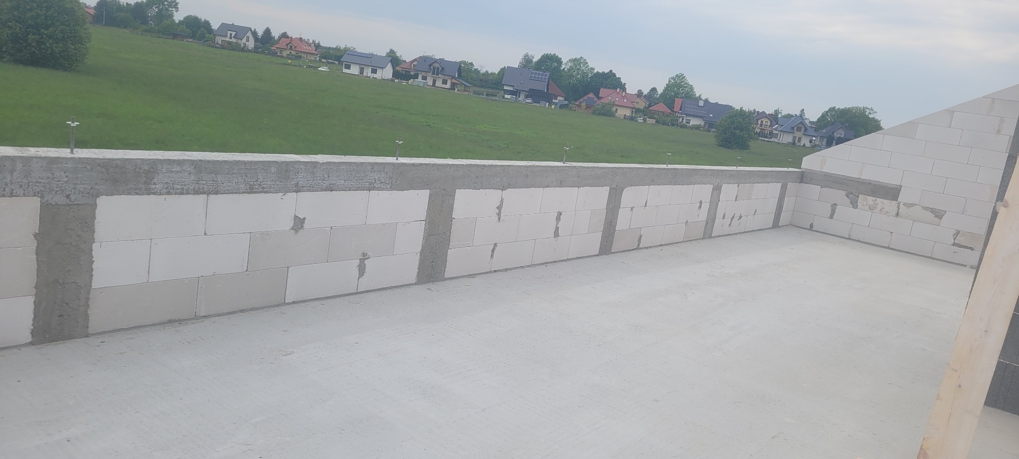 Ściana z białych bloczków z betonu komórkowego w trakcie budowy domu, widoczne spoiny i zbrojenie, w tle zielone wzgórze z zabudową.