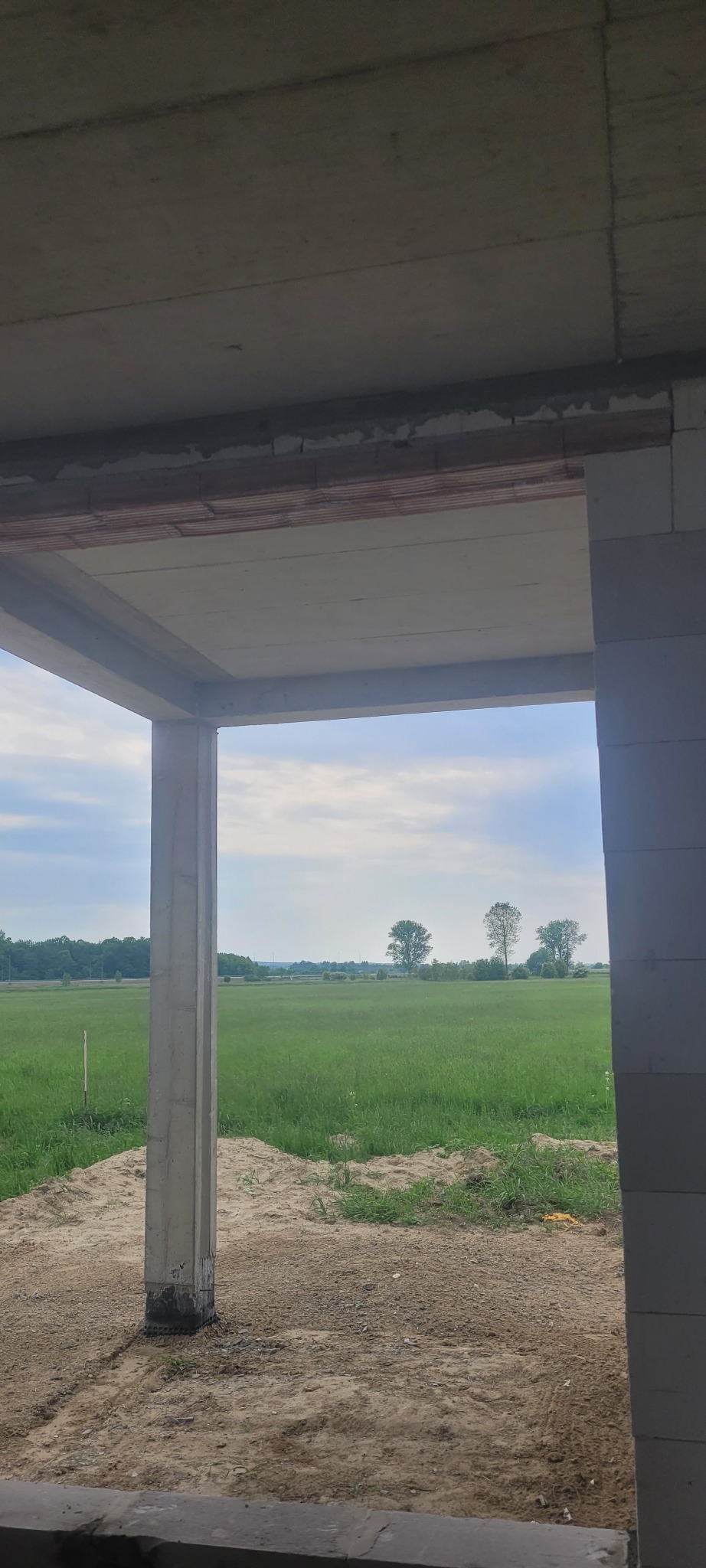 Surowy stan domu murowanego z widokiem na zielone pole i drzewa przez otwór okienny, widoczne bloczki betonowe i nadproże.