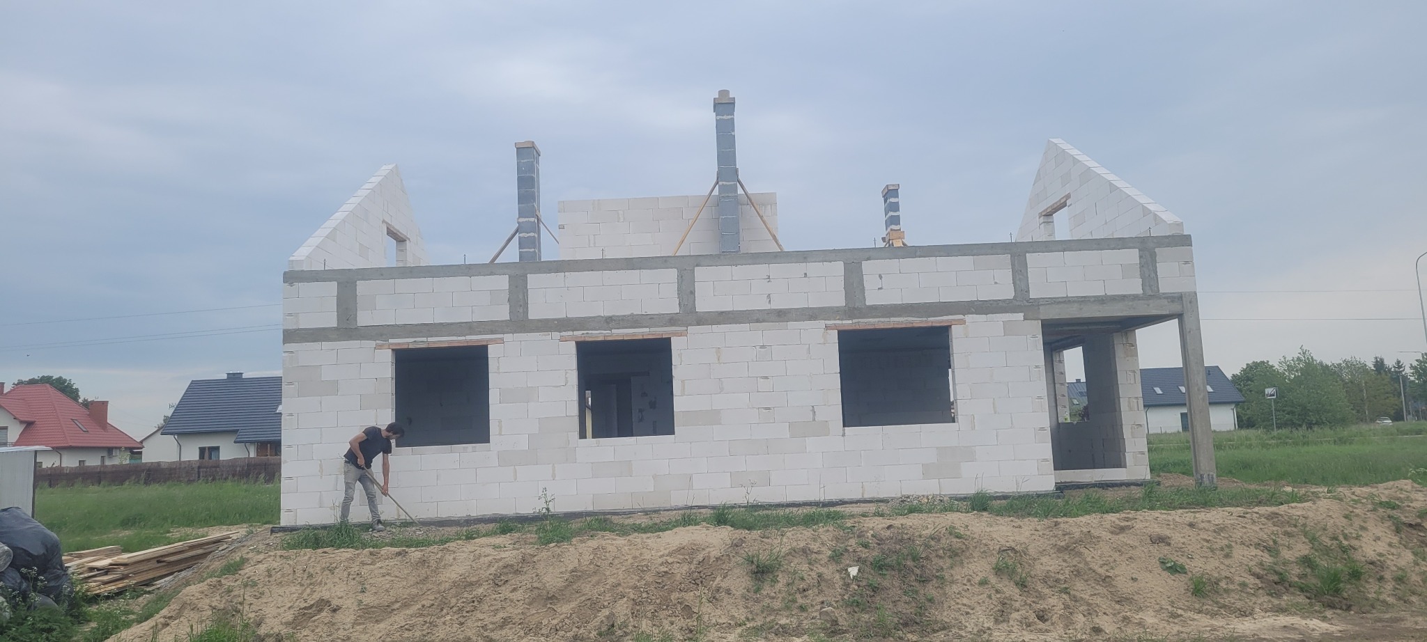 Wznoszony dom z białych bloczków, widoczne otwory okienne i kominy, pracownik wyrównuje teren przed budynkiem.