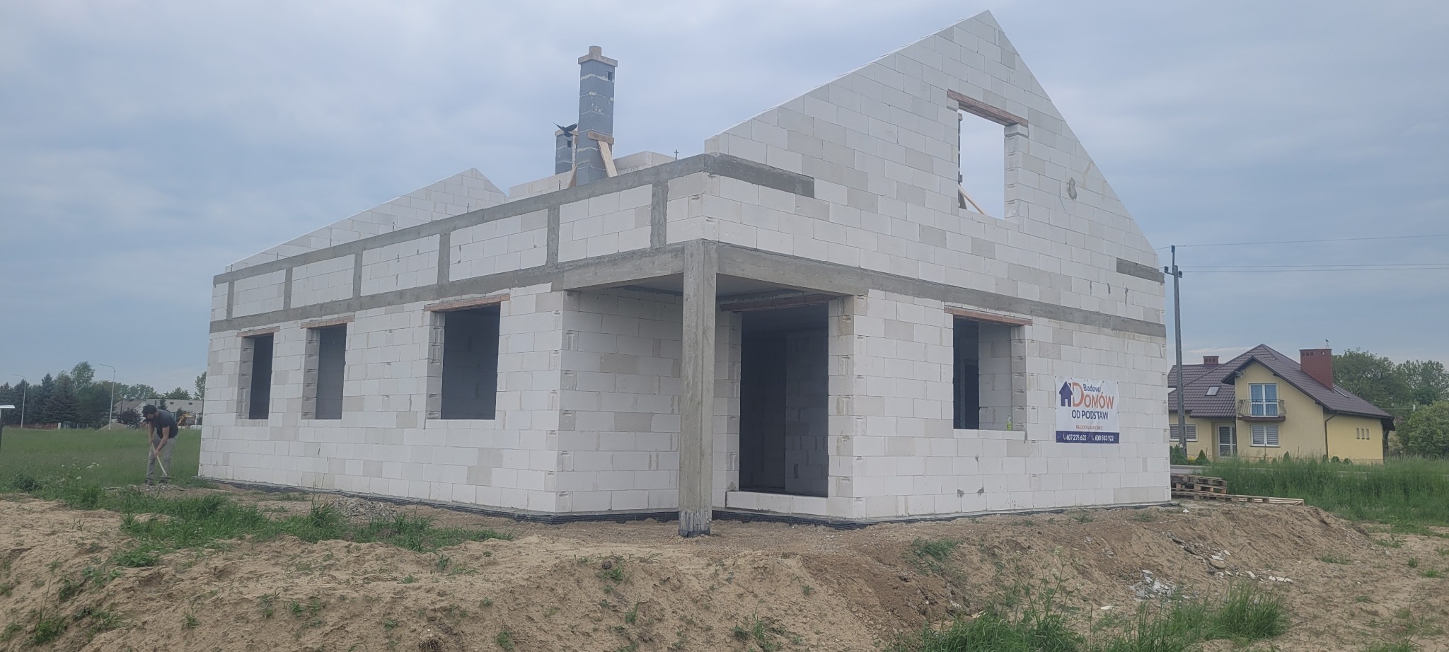 Surowy stan domu z białych bloczków, widoczne nadproża, kominy w trakcie budowy, na pierwszym planie ziemia, w tle dom jednorodzinny oraz pracownik z łopatą.