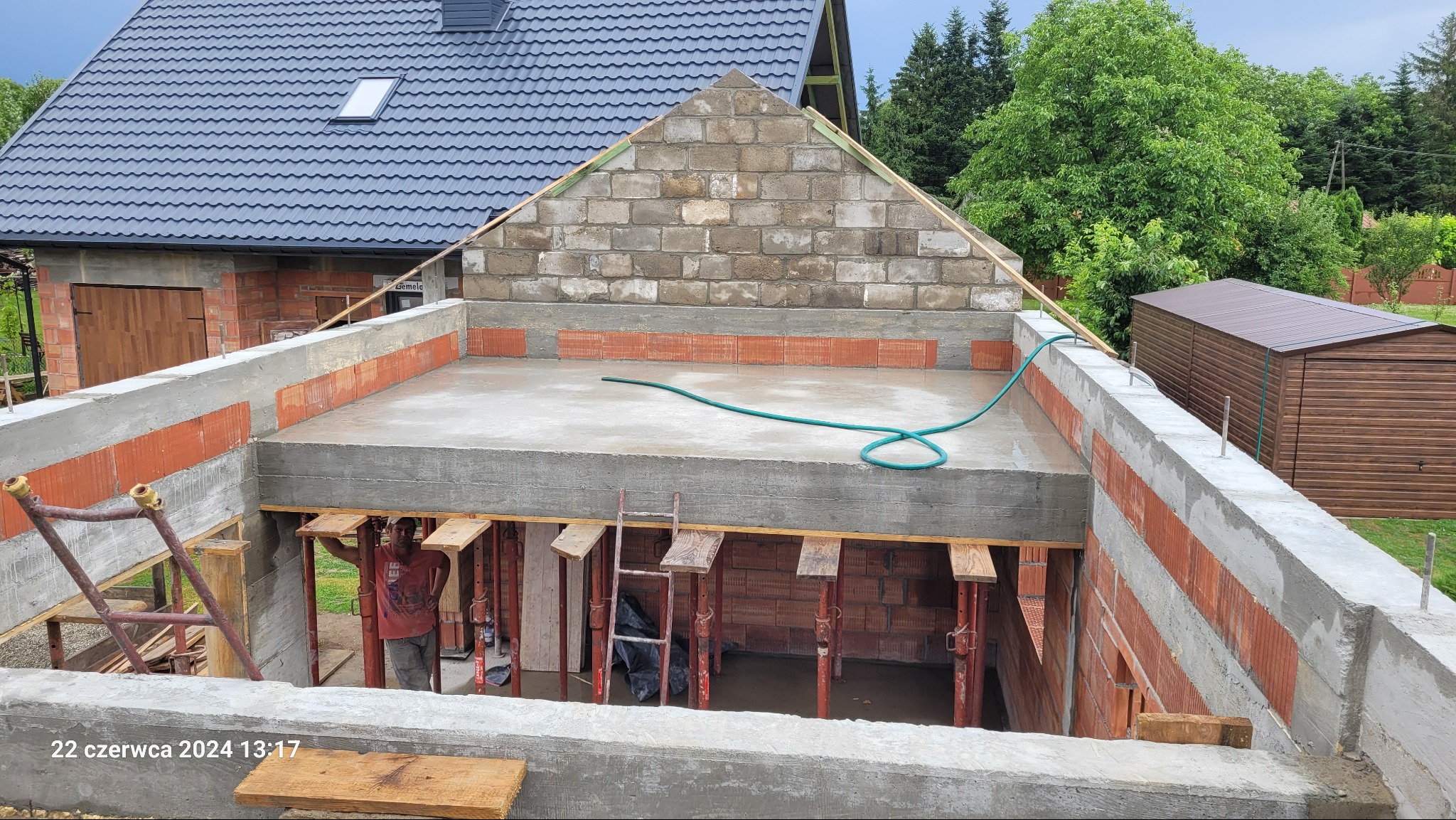 Budowa domu w trakcie realizacji: wylany betonowy strop, ściany z czerwonej cegły, widoczne podpory i szalunki, fragment murowanej ściany szczytowej z bloczków betonowych, zielony wąż na stropie.