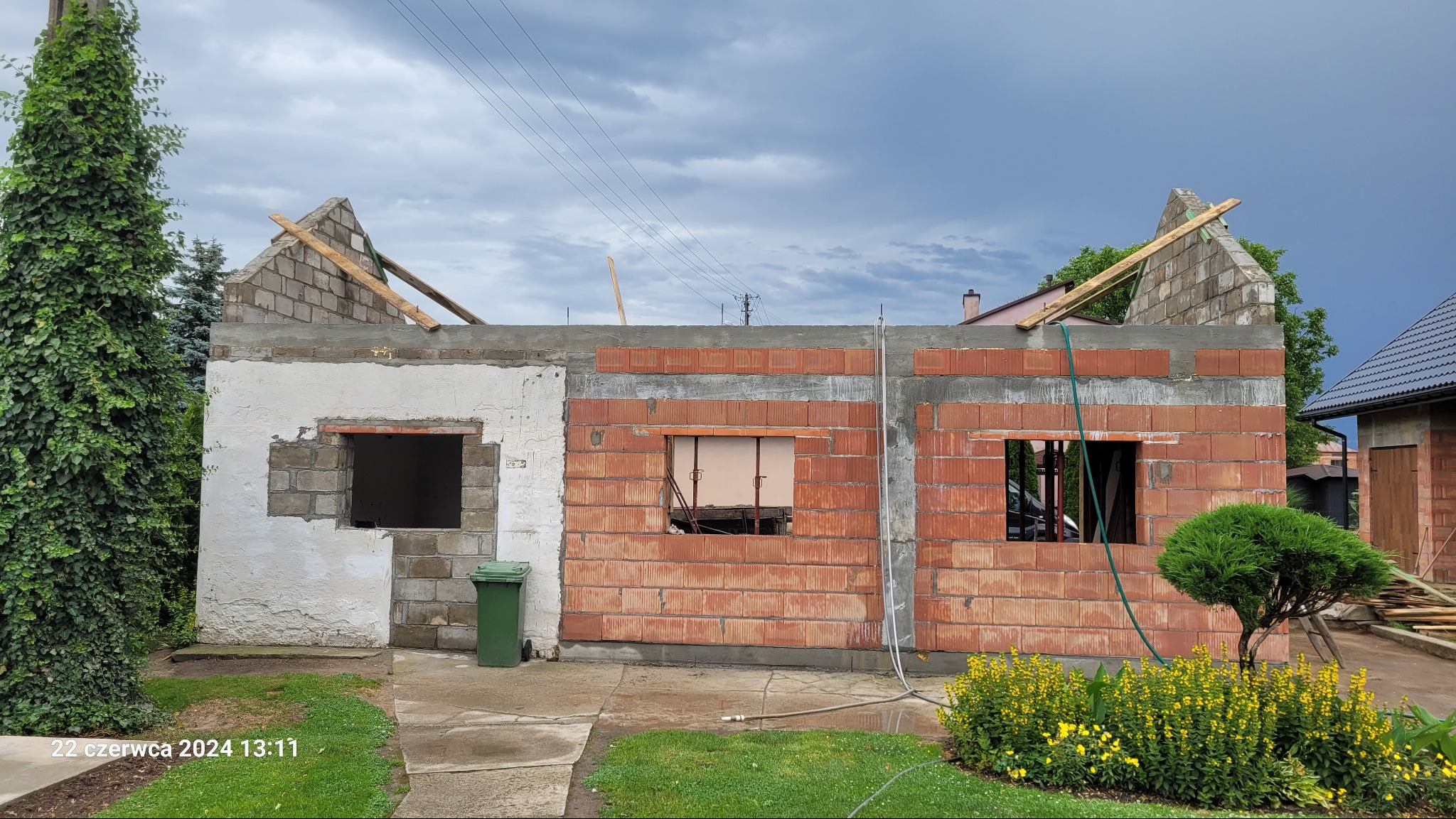 Budowa domu w trakcie: ściany z czerwonej cegły i szarego betonu, widoczne otwory okienne, częściowo otynkowana ściana z pustaków, zielony krzew na pierwszym planie, pochmurne niebo w tle.