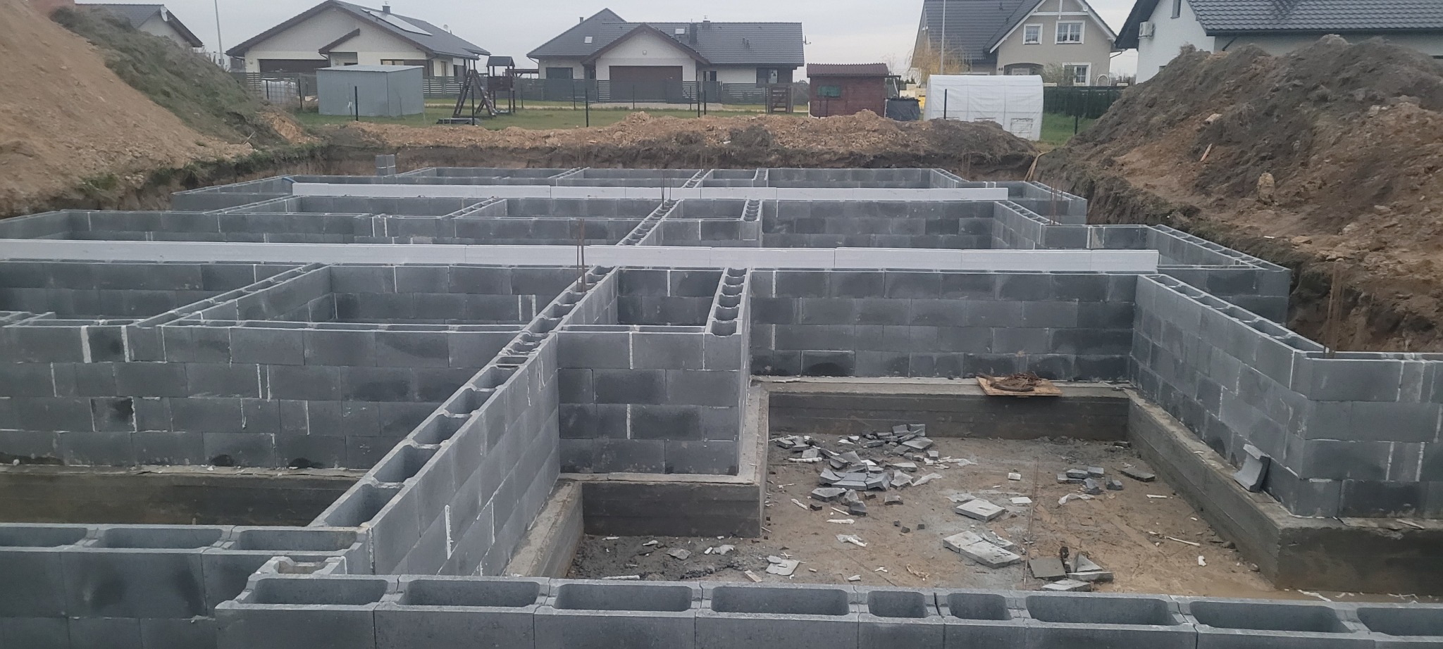 Fundamenty z szarych bloczków betonowych w wykopie, widoczne zbrojenia pionowe, w tle domy jednorodzinne w zabudowie szeregowej.