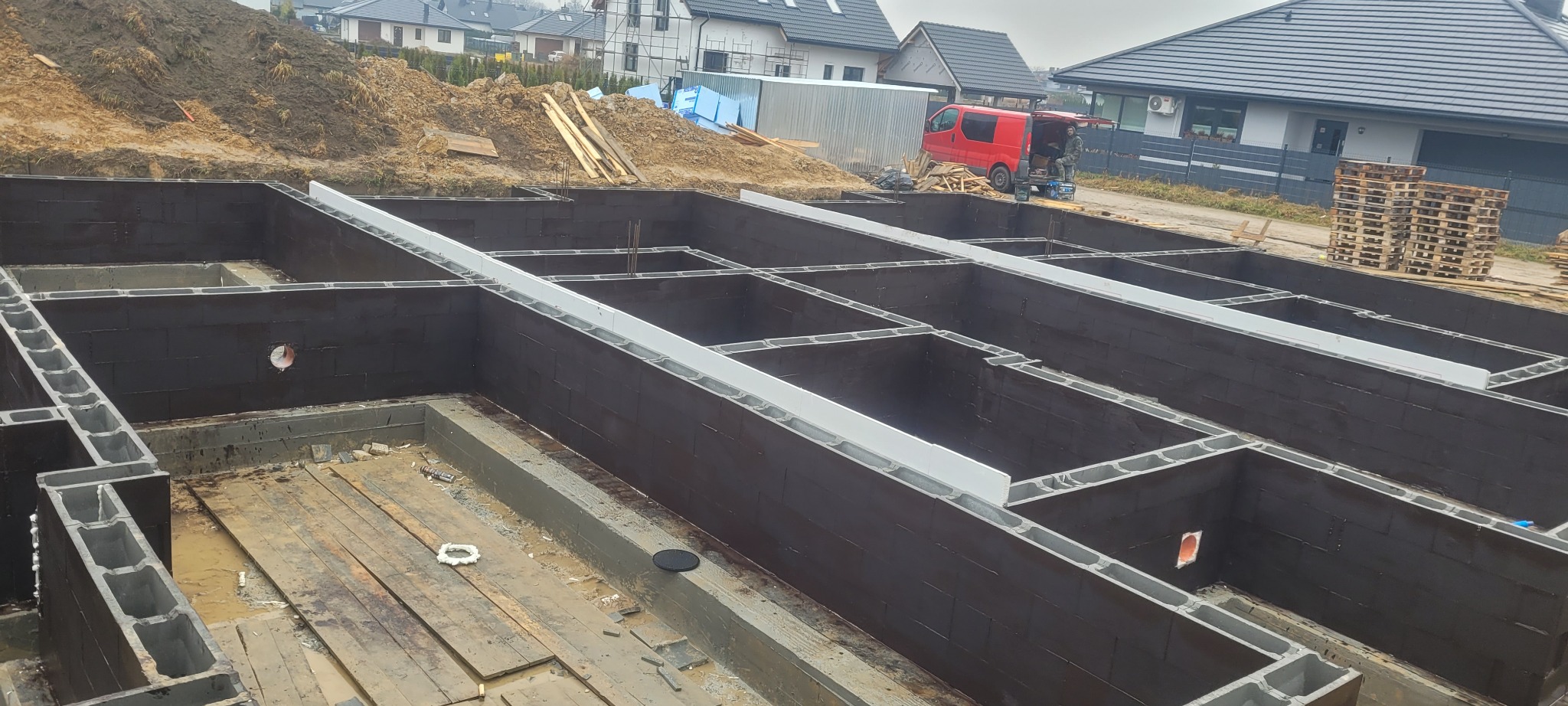 Zabezpieczone fundamenty z bloczków betonowych pokryte czarną izolacją, z widocznymi szalunkami i stertą ziemi w tle, na placu budowy z czerwonym samochodem dostawczym w oddali.
