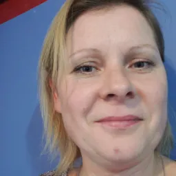 Portret uśmiechniętej kobiety z blond włosami, na tle niebieskiej ściany z czerwonym elementem konstrukcyjnym u góry.