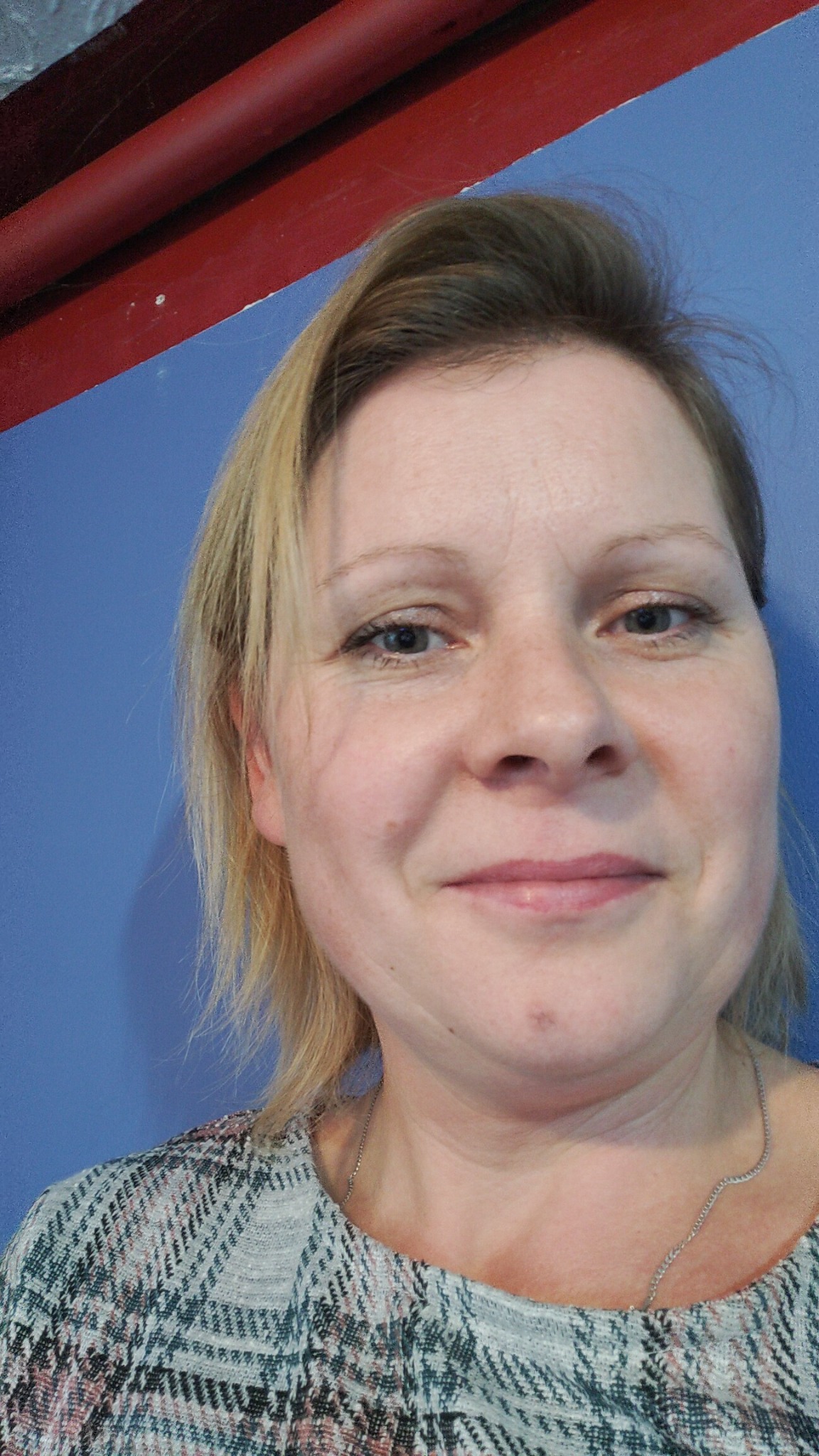 Portret uśmiechniętej kobiety z blond włosami, na tle niebieskiej ściany z czerwonym elementem konstrukcyjnym u góry.