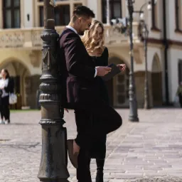 Elegancka para w garniturze i sukience przegląda tablet oparta o starą latarnię na brukowanym placu miejskim.