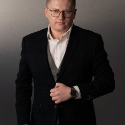 Opaliński Nieruchomości - Portret mężczyzny w okularach, czarnym garniturze i kamizelce w kratę, z zegarkiem na ręce, pozującego na szarym tle.