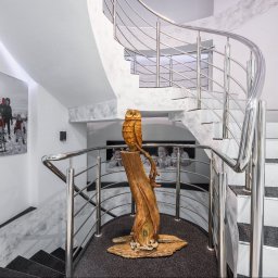 Opaliński Nieruchomości - Nowoczesna klatka schodowa z spiralnymi schodami, stalowymi balustradami, drewnianą rzeźbą sowy na postumencie i obrazem z motywem zimowym na ścianie.
