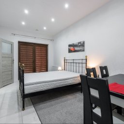 Opaliński Nieruchomości - Jasne wnętrze pokoju z czarnym łóżkiem z metalową ramą, stolikiem z krzesłami i telewizorem na ścianie; na ścianie wisi obraz z motywem Paryża.